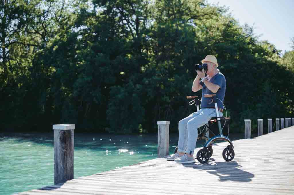 A man uses a camera to photograph the sea from a jetty while sitting on a Saljol walking frame