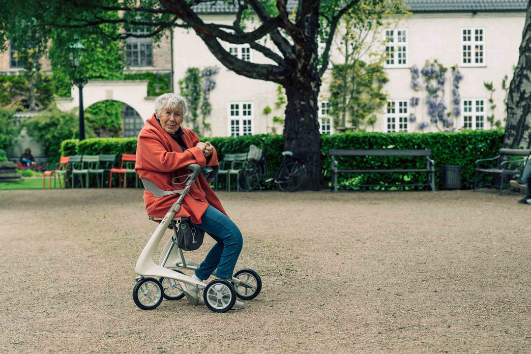 A woman sits on a 'byACRE Carbon Ultralight' walker in a cobble stone court yard