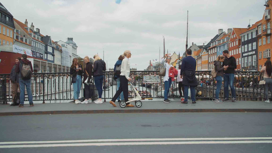 A woman crosses over a bridge in Copenhagen with a 'byACRE Carbon Ultralight' rollator. 