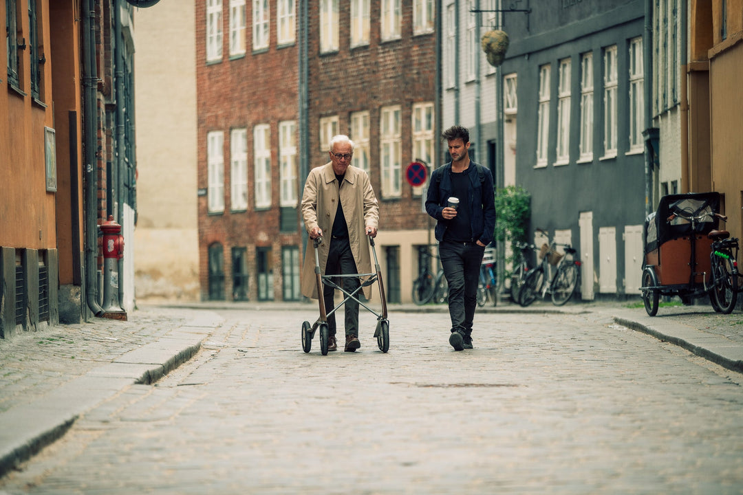 A man walks with the assistance of a 'byACRE Overland' rollator frame, side by side with his friend, along a street in Copenhagen