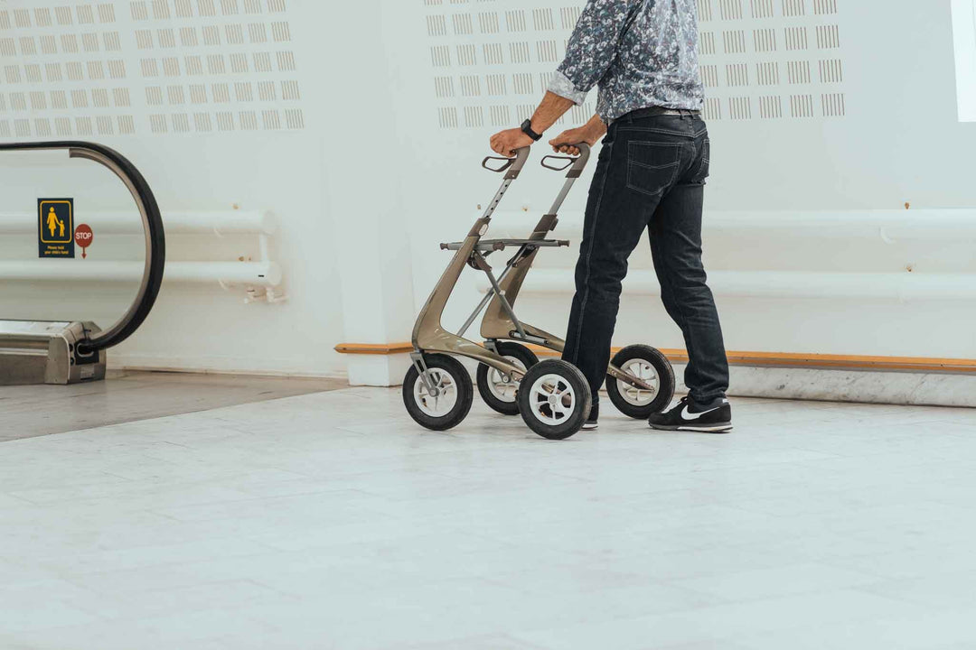 A person uses a 'byACRE Carbon Overland' walking frame as they walk towards an escalator. 