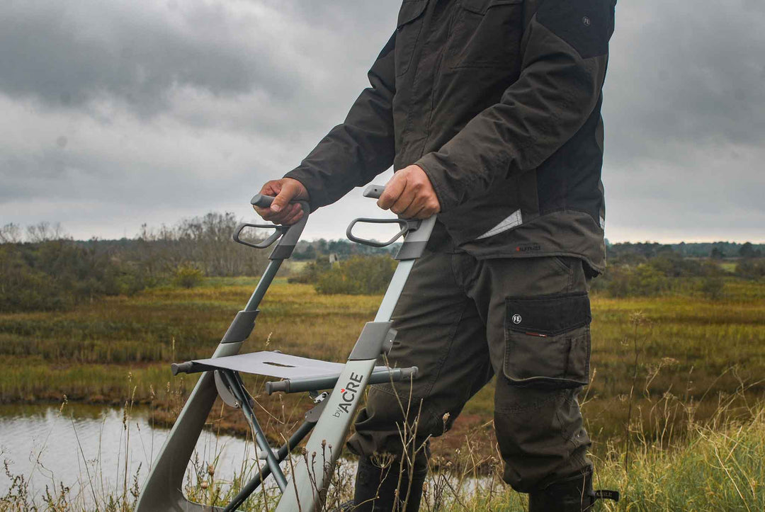A close of the handles of a 'byACRE Carbon Overland' walking frame being used by a man walking in a field beside a swamp