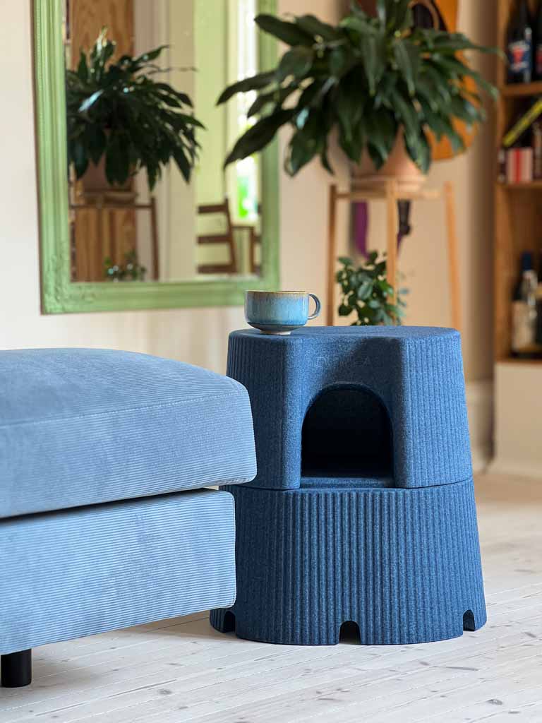 The blue Dorica multifunctional stool placed beside a couch with a cup on top and a plant in the background