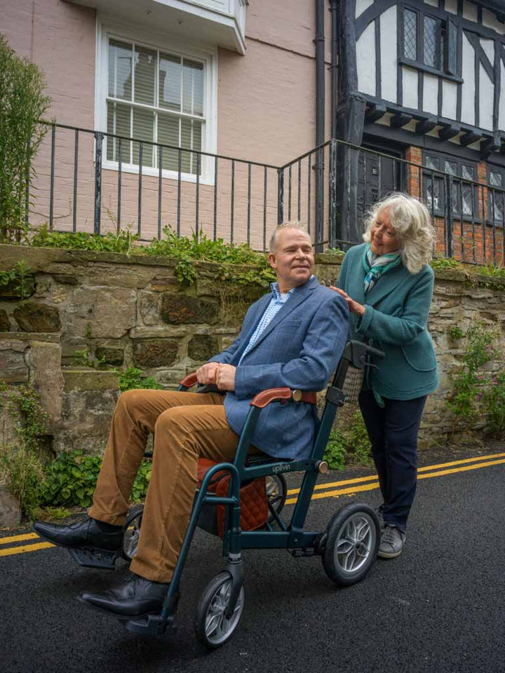 Man in a wheelchair being pushed by a woman on a street with a house in the background