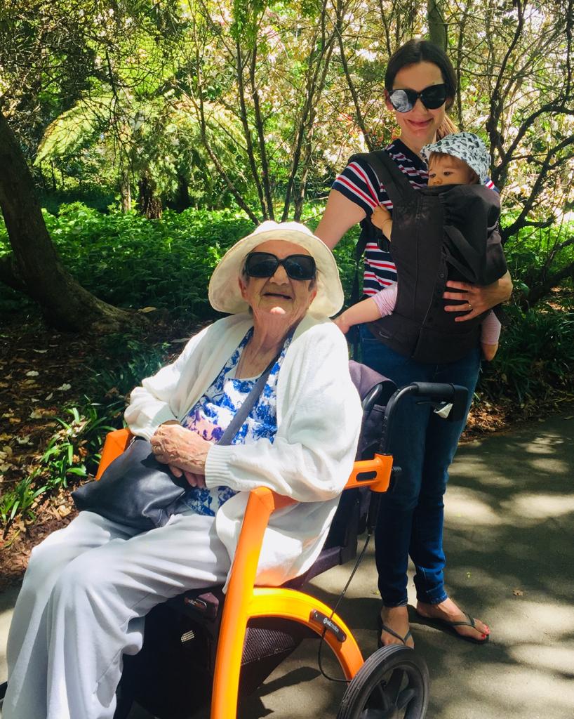 A woman in a striped shirt and sunglasses carries a baby in a black baby carrier while standing next to an elderly woman sitting in an orange  wheelchair in a sunny park.