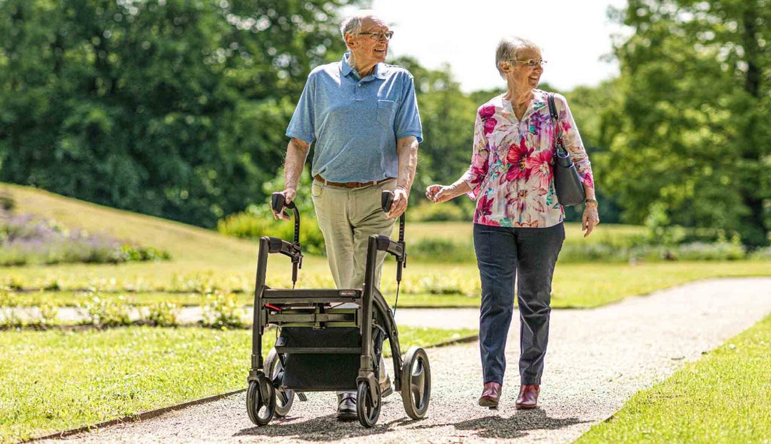 An elderly man walks with a 'Rollz Motion' walking frame that is deisgned for people with Parkinson's disease. He is accompanied by his wife, they are both smiling, strolling in the park with trees in the background.