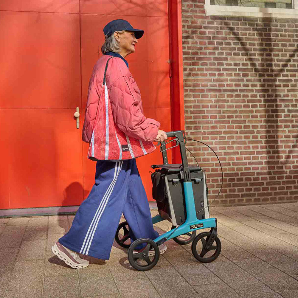 women walking with a blue rollator on a footpath beside a red door and brick wall.