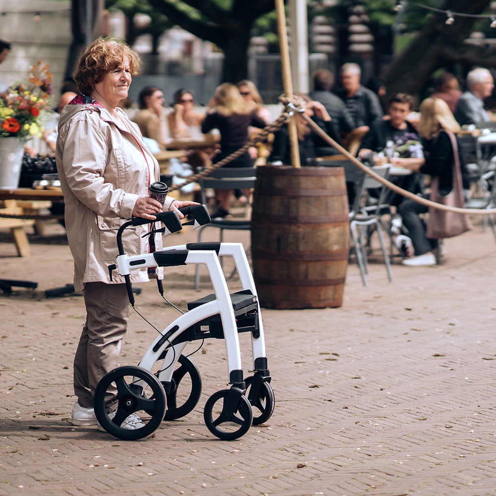 A woman stands with a 'Rollz Motion' walking frame in a town square with outdoor restaurant seating in the background