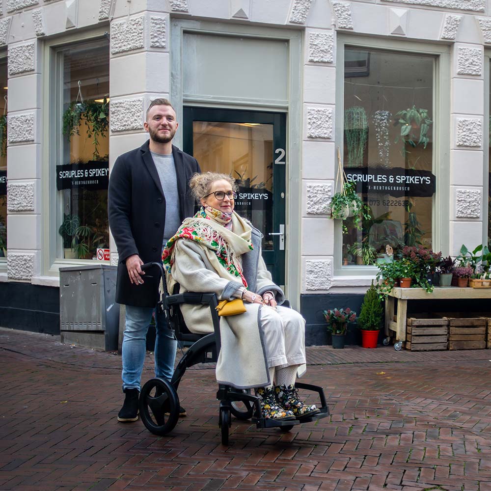 Man pushing a woman in a wheelchair on a street with a building and storefront in the background.