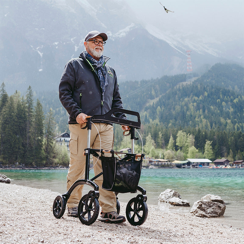 Man with a walker standing by a lake with mountains in the background