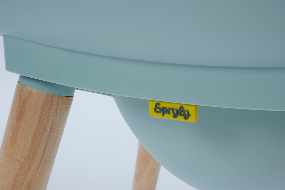 Close-up of a light blue Spryly commode  showing the underside of the seat, a small yellow brand label, and part of a natural wood leg.