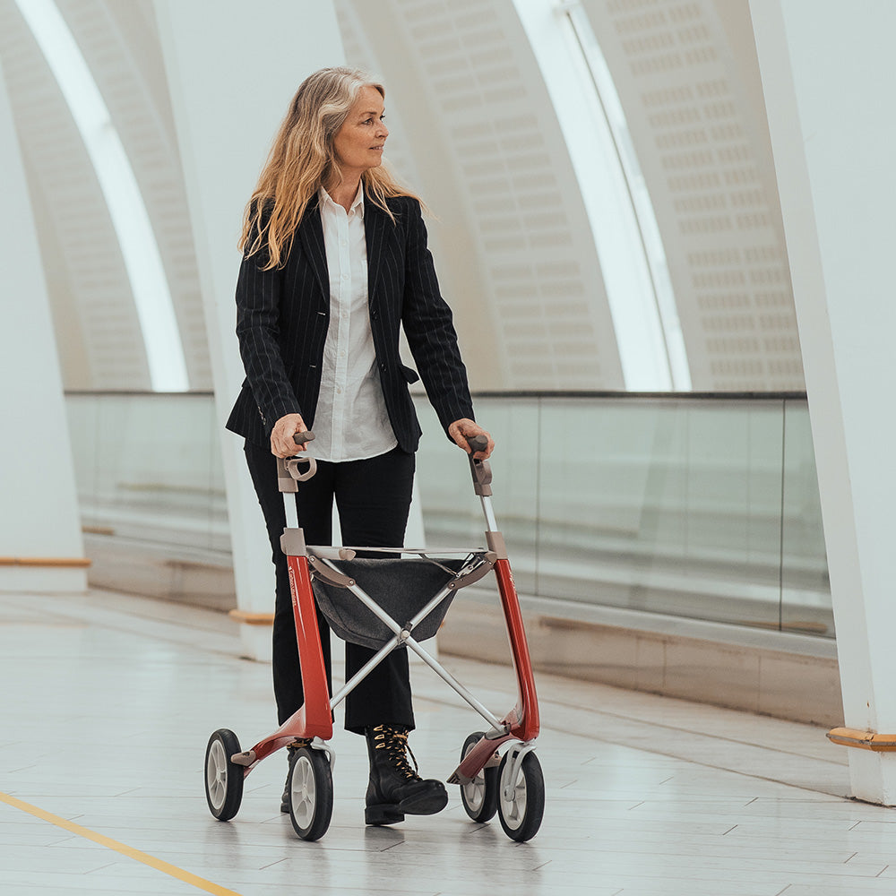 A trendy woman dressed in boots and a suit walks with a modern lightweight walker that is red, made by byACRE, inside a train station with white tiles.