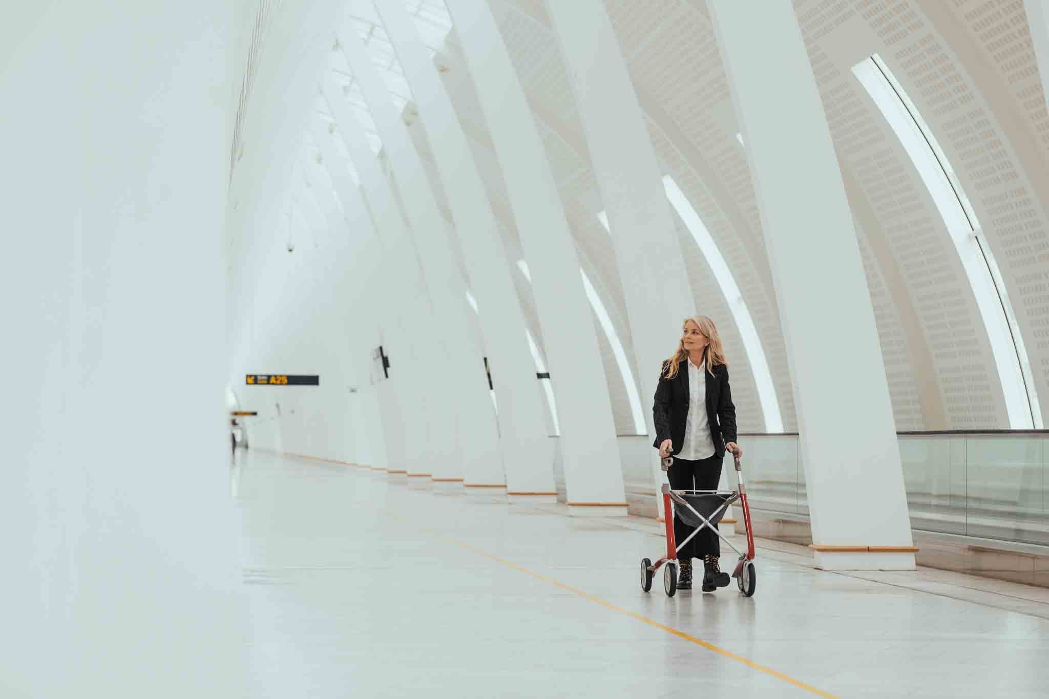 A woman walks with a modern lightweight 'byACRE Ultralight' walking frame inside a modern train station with white arches.