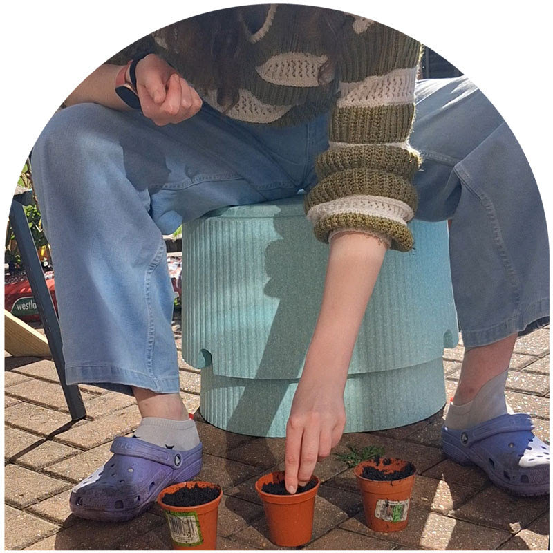 Person sitting on a green stool planting small plants in orange pots on a sunny day.