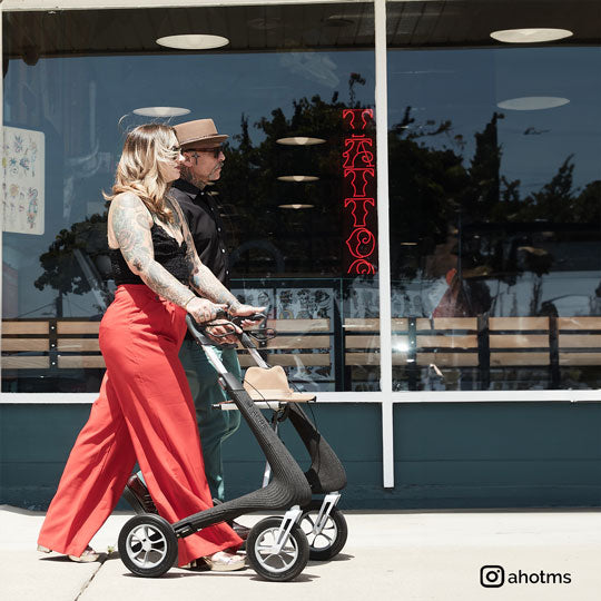 A woman walks along a footpath with a fashionable looking walker rollator with a tattoo parlor in the background