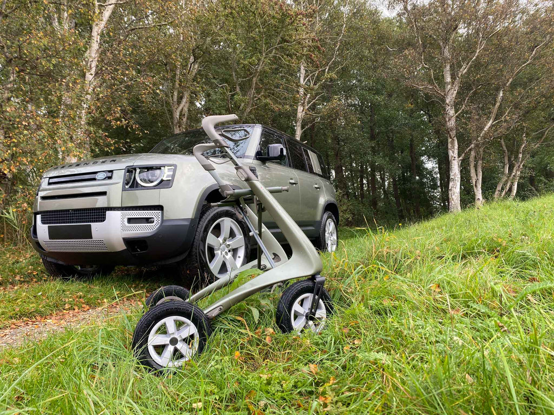 A 'byACRE Overland' walking frame is positioned in front of a 'Land Rover Defender' car, in a grassy field with trees in the background