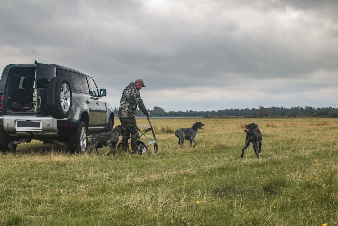 A man walks away from his SUV into a field using a 'byACRE Carbon Overland' walking frame, accompanied by three friendly dogs