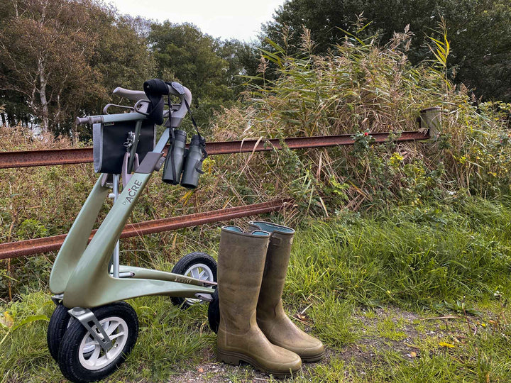 A 'byACRE Carbon Overland' walking frame is folded and stands beside a pair of brown gumboots on a background of long grass and an old fence.